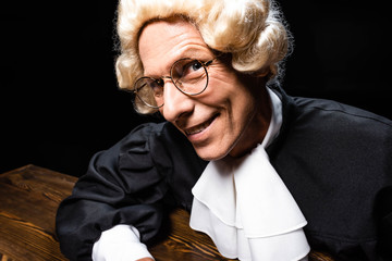 smiling judge in judicial robe and wig sitting at table isolated on black