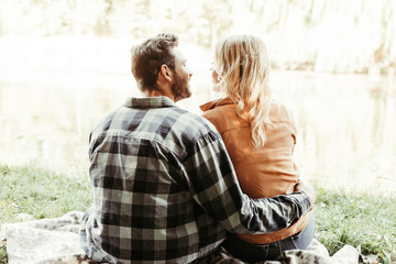 back view of young man embracing girlfriend while sitting near lake in park