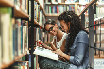 Two female students reads a book by the bookshelf at the library. Reading a book.	
