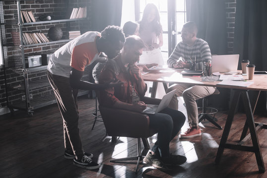 young african american businessman standing near thoughtful colleague sitting in armchair and using laptop