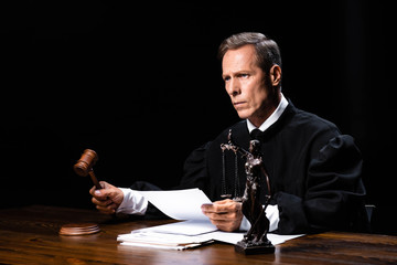 judge in judicial robe sitting at table and holding paper and gavel isolated on black