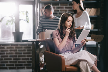 Fototapeta premium thoughtful businesswoman sitting in armchair and using digital tablet near young colleagues