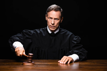 judge in judicial robe sitting at table and hitting with gavel isolated on black