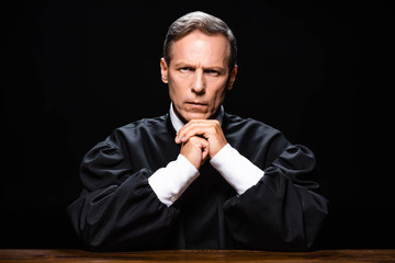 judge in judicial robe sitting at table and looking at camera isolated on black © LIGHTFIELD STUDIOS