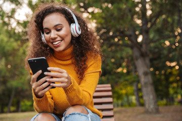 Curly woman sit on bench in park using mobile phone.