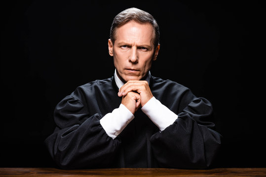 judge in judicial robe sitting at table and looking at camera isolated on black