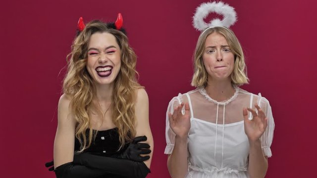 Happy demon girl incites to action and the sad angel girl dissuades them in carnival costumes isolated over red wall background