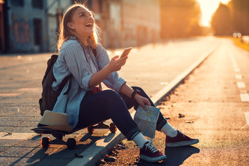 Smiling girl sitting on long board in the city during sunset and looking at phone.