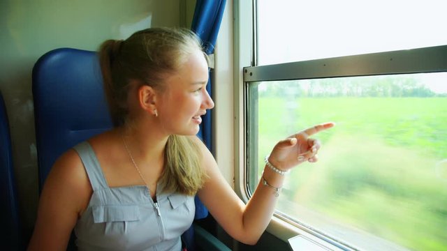cute girl with long hair in ponytail in summer dress looks out window in intercity train laughs pointing to landscape
