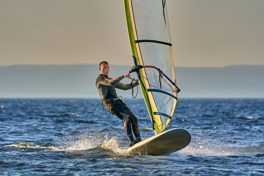 A male athlete is interested in windsurfing. He moves on a Sailboard on a large lake on an autumn day.