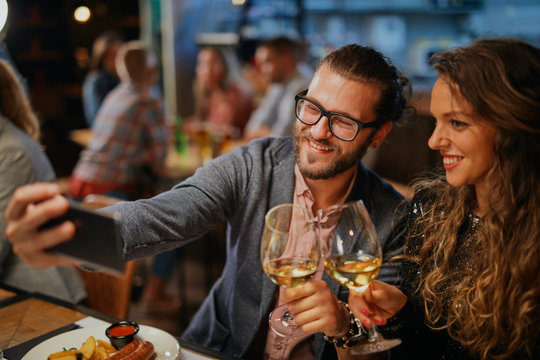 Cute caucasian fashionable couple sitting in restaurant at dinner holding glasses of wine and taking selfie.