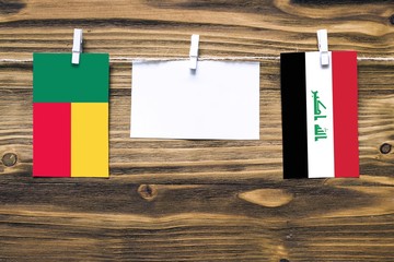 Hanging flags of Benin and Iraq attached to rope with clothes pins with copy space on white note paper on wooden background.Diplomatic relations between countries.