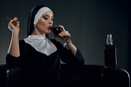 Cropped half-turn shot of a nun, sitting on a chair. She's wearing dark nun's clothing. The nun is drinking wine and holding cigarette in her hand. There is a bottle with alcohol on a chair armrest. 