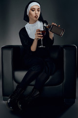 Full length shot of a nun, sitting on a chair. She's wearing dark nun's clothing and big boots. The nun is smoking, holding glass of wine in right hand and pour drink into glass. 