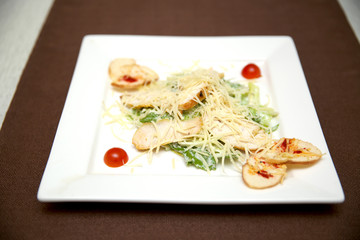 Caesar salad on a square white plate. square dish of lettuce on a table with a brown tablecloth. salad with greens, grated cheese and cherry tomatoes beautifully served.