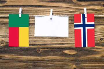 Hanging flags of Benin and Norway attached to rope with clothes pins with copy space on white note paper on wooden background.Diplomatic relations between countries.