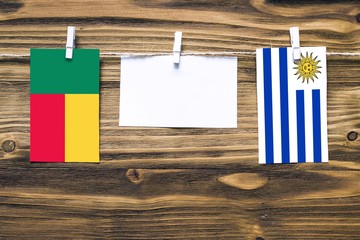 Hanging flags of Benin and Uruguay attached to rope with clothes pins with copy space on white note paper on wooden background.Diplomatic relations between countries.