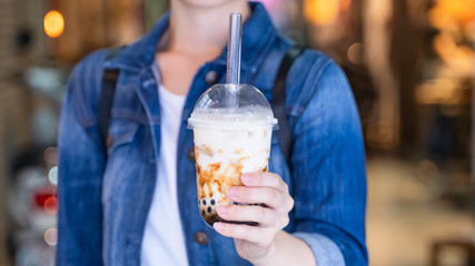 Young woman is holding, drinking brown sugar flavored tapioca pearl bubble milk tea with glass straw in night market of Taiwan, close up, bokeh