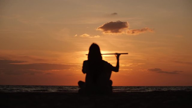 Silhouette of a beautiful man with long hair playing on flute in front of sunset. Slow motion. Enlightenment