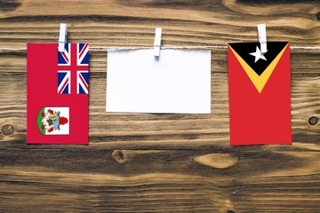 Hanging flags of Bermuda and East Timor attached to rope with clothes pins with copy space on white note paper on wooden background.Diplomatic relations between countries.