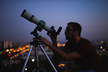 Astronomer with a telescope watching at the stars and Moon with blurred city lights in the...
