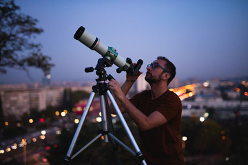 Astronomer with a telescope watching at the stars and Moon with blurred city lights in the background.