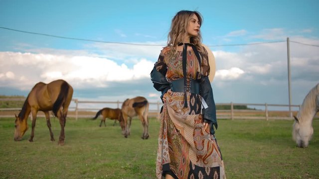 Young female in ornamental dress and stylish jacket posing for camera while spending time in ranch enclosure near horses