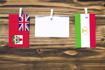 Hanging flags of Bermuda and Tajikistan attached to rope with clothes pins with copy space on white note paper on wooden background.Diplomatic relations between countries.