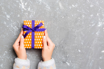 Female's hands holding striped gift box with colored ribbon on gray cement background. Christmas...