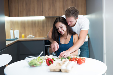 Obraz na płótnie Canvas young couple cooking breakfast in the kitchen husband stands behind and hugs his beloved wife while they cut vegetables together family traditions