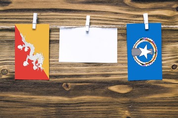 Hanging flags of Bhutan and Northern Mariana Islands attached to rope with clothes pins with copy space on white note paper on wooden background.Diplomatic relations between countries.