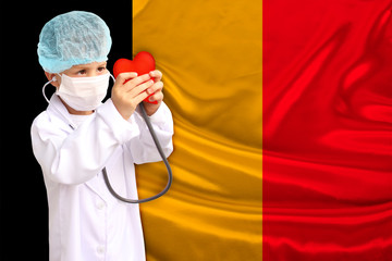 child, boy, in a white doctor’s coat, hat and mask attached a stethoscope to a red heart model,...