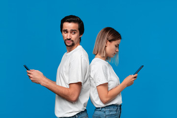 Blond woman and brunet man in white tees and blue jeans with mobile phones standing isolated over blue background. Man tries to see what is on a screen of his freind.