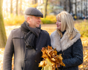 Happy blonde mature woman and handsome middle-aged brunette man walk in park, looking at each other. Loving couple of 45-50 years old walks in autumn park in warm clothes, holding bouquet of leaves