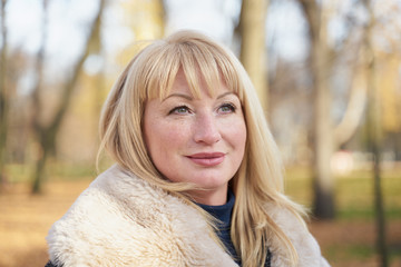 Closeup portrait of happy blonde mature woman is standing in autumn park and thinking. Beautiful woman is relaxing in nature on sunny day. Portrait of middle aged woman smiling and daydreaming.