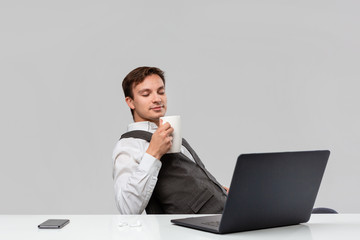 Businessman in a white t-shirt and grey vest drinking coffee sitting at the white table with laptop and smartphone.