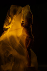 Body of a beautiful girl gracefully fits a cellophane film fluttering in the wind. In defocusing, it is illuminated with a fiery yellow color. Artistic, conceptual, avant-garde photography.