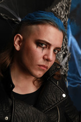 Teen alt boy with blue hair and shaved head dressed in black on dark background