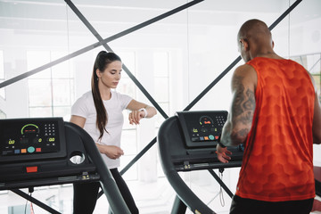 attentive trainer looking at fitness tracker while standing near african american sportsman running on treadmill