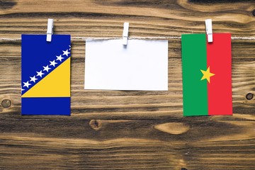 Hanging flags of Bosnia Herzegovina and Burkina Faso attached to rope with clothes pins with copy space on white note paper on wooden background.Diplomatic relations between countries.