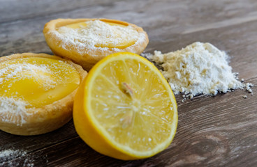 Close-up view of freshly made lemon cakes shown on a traditional wooden table together with a sliced lemon and baking flour used in the bakin