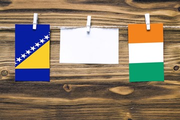 Hanging flags of Bosnia Herzegovina and Cote D'Ivoire attached to rope with clothes pins with copy space on white note paper on wooden background.Diplomatic relations between countries.