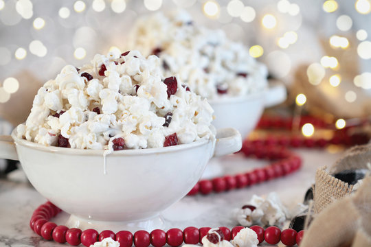Bowls of homemade popcorn and dried cranberry snack covered in white chocolate ready for the holidays surrounded by bokeh lights and red bead garland.. Selective focus with blurred background