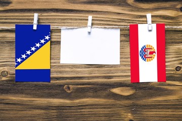 Hanging flags of Bosnia Herzegovina and French Polynesia attached to rope with clothes pins with copy space on white note paper on wooden background.Diplomatic relations between countries.