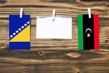 Hanging flags of Bosnia Herzegovina and Libya attached to rope with clothes pins with copy space on white note paper on wooden background.Diplomatic relations between countries.