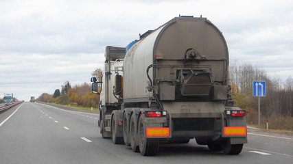 Bitumen truck with a round barrel semi trailer moving on a two-lane asphalted country road in autumn day, Logistics, hot liquid material road transportation, rear view