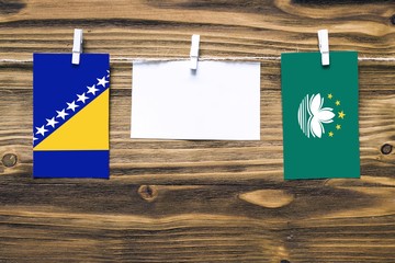 Hanging flags of Bosnia Herzegovina and Macao attached to rope with clothes pins with copy space on white note paper on wooden background.Diplomatic relations between countries.