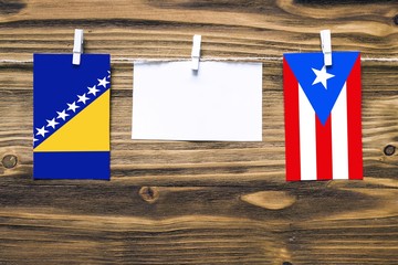 Hanging flags of Bosnia Herzegovina and Puerto Rico attached to rope with clothes pins with copy space on white note paper on wooden background.Diplomatic relations between countries.