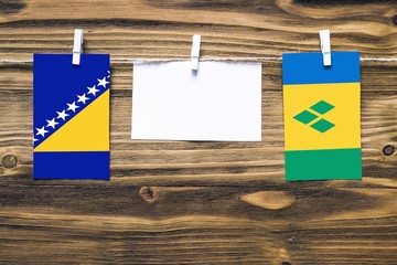 Hanging flags of Bosnia Herzegovina and Saint Vincent And The Grenadines attached to rope with clothes pins with copy space on white note paper on wooden background.Diplomatic relations.