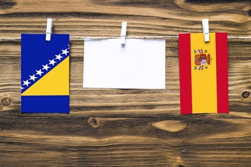 Hanging flags of Bosnia Herzegovina and Spain attached to rope with clothes pins with copy space on white note paper on wooden background.Diplomatic relations between countries.